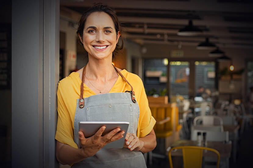 Vrouw met schort aan en digitaal apparaatje in handen leunt tegen de deurpost. Ze kijkt lachend de camera in. Op de achtergrond de setting van een restaurant.