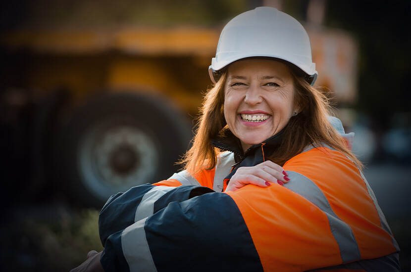 Vrouw met oranje veiligheidsjas en witte helm zit met haar handen over elkaar en kijkt lachend de camera in.