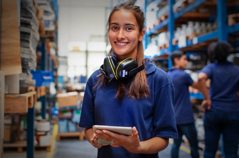 Vrouw met een blauw shirt en gehoorbescherming om haar hals heeft een i-pad in handen en kijkt lachend de camera in. Ze staat in de omgeving van een magazijn.